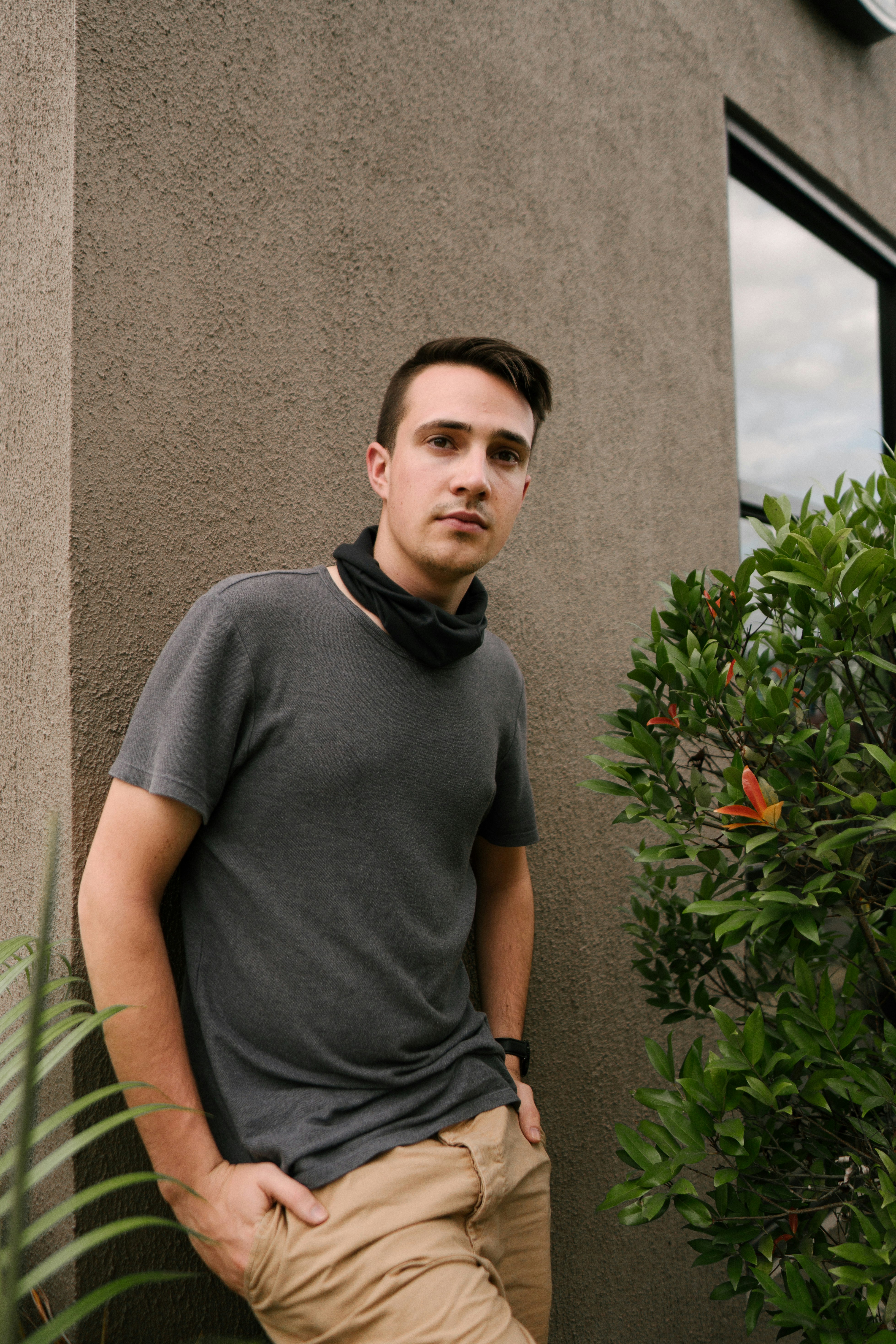 man standing with both hands in his bottoms's pockets while leaning back on wall and looking at the camera during day